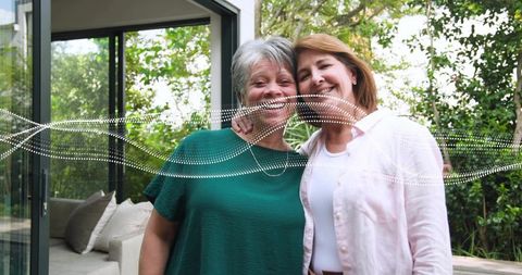 Smiling mother and adult daughter hugging on patio with garden view and warm sunlight