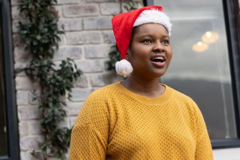 African american woman wearing santa hat and mustard sweater singing outdoors, festive portrait