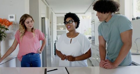 Image of a young couple in casual attire engaging with a professional real estate agent in a bright, modern kitchen setting. They appear to be democratically involved in a consultation, possibly discussing property features and potential purchase. The scene conveys a sense of trust, collaboration, and commitment to finding the right home. Ideal for use in real estate marketing materials, property websites, or articles related to home buying processes and consultations.