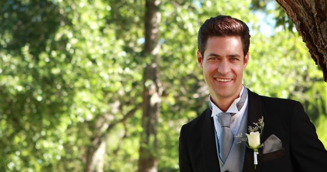 Depicts a groom smiling in formal wedding attire, posing against a natural outdoor backdrop with lush greenery. Ideal for use in wedding magazines, event planning brochures, romantic greeting cards, or websites focusing on wedding services and celebrations.