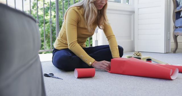 Woman is sitting on beige carpet, wrapping gift with red wrapping paper. Scissors and other wrapping accessories are within reach. Suitable for illustrations of holiday preparations, gift-giving, home activities, and festive celebrations.