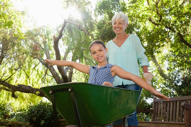 Smiling senior woman pushing granddaughter sitting in wheelbarrow at backyard