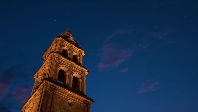 Illuminated baroque bell tower at twilight with cross and bells against deep blue sky