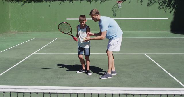 A father teaching his young son to play tennis on an outdoor court. They are both standing on the court, with the father helping the son with his grip on the racket. The scene is bright and sunny, indicating a warm summer day. This can be used for marketing family sports programs, promoting outdoor activities, or illustrating articles about family bonding and sports coaching.