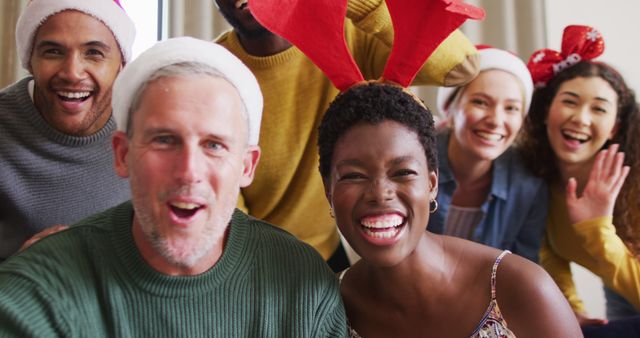 This image shows a group of friends with diverse backgrounds celebrating Christmas together indoors. They are all smiling and wearing festive accessories such as Santa hats and an antler headband, creating a joyful atmosphere. This is ideal for content relating to holiday celebrations, festive spirit, multicultural gatherings, and friendship.