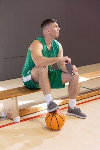 Male basketball player taking break on bench with foot on ball holding water bottle