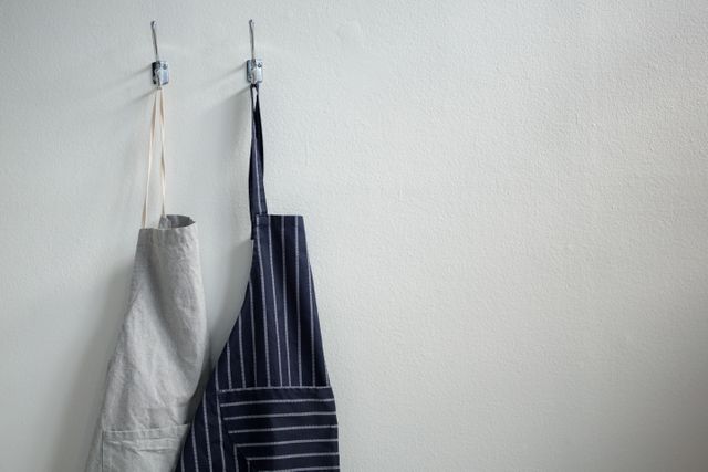 Two aprons hanging on metal hooks against a plain wall. One apron is plain gray and the other is black with white stripes. Ideal for use in articles or advertisements related to cooking, home organization, kitchen decor, or minimalist living.