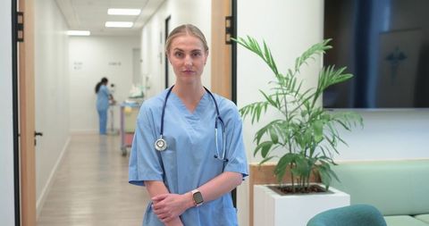 Confident female nurse standing in clinic corridor wearing blue scrubs and stethoscope