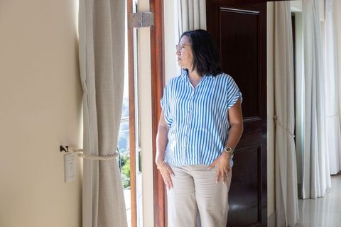 Mature asian woman gazing through balcony door in striped blouse and glasses, sunlit interior