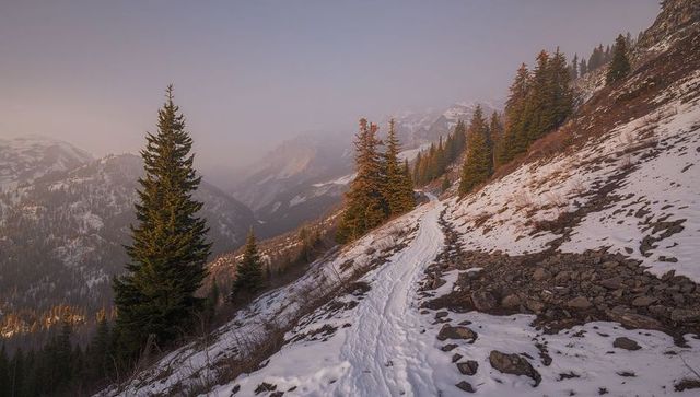 Winding snow-covered mountain trail climbing alpine ridge through evergreen forest at dusk