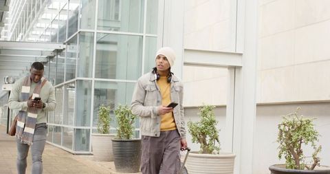 Diverse young men walking past glass facade pulling suitcase and checking smartphones