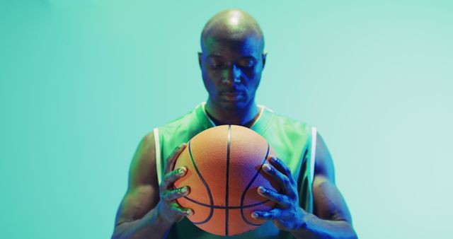 Depicting a male athlete concentrating while holding a basketball, this image captures the determination and focus inherent in sports. The neon green light adds a modern, dramatic effect, perfect for illustrating themes of sportsmanship, perseverance, and athletic training for sports magazines, ads, and promotional materials.