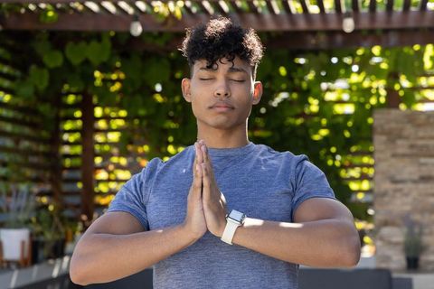 Young african american man meditating under pergola in sunlit backyard wearing smartwatch