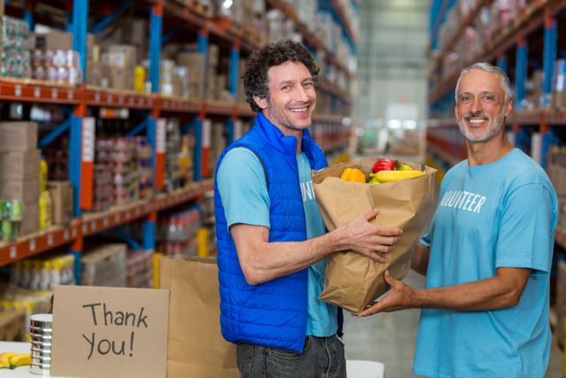 Two happy volunteers holding a grocery bag filled with fruits and vegetables in a warehouse. They are smiling and wearing blue shirts, indicating their involvement in community service. Shelves filled with boxes and a thank you sign are visible in the background. Ideal for use in articles or campaigns promoting volunteer work, charity events, food donation drives, and community service initiatives.