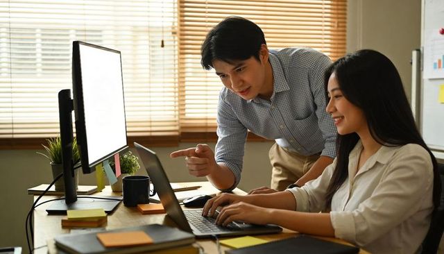Colleagues collaborating at desk, mentor pointing at laptop while teammate typing
