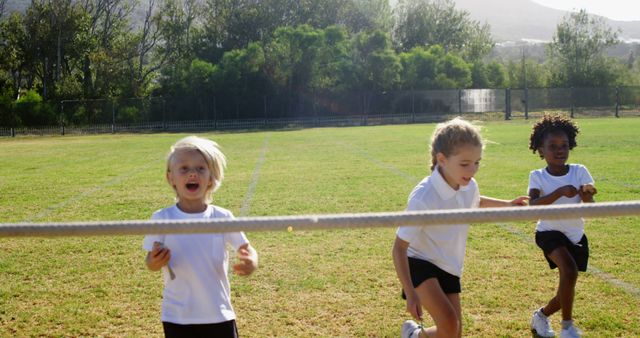 Three young children participating in a sports day race on a sunny day. They are wearing white T-shirts and black shorts and are visibly energetic and enthusiastic. The outdoor setting is a green grass field with trees in the background, suggesting a community or school sports event. Ideal for use in educational materials, fitness campaigns, children's activities, and family-oriented promotions.