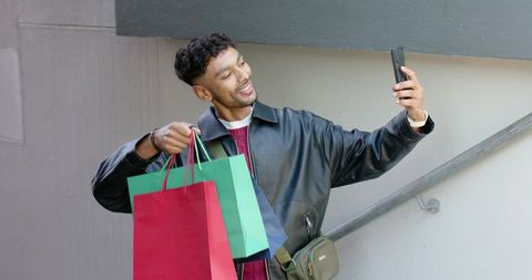 Man taking selfie with colorful shopping bags on stair landing wearing leather jacket