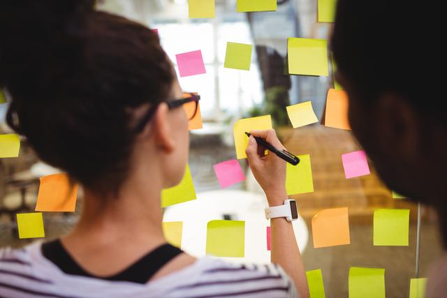 Business executives are brainstorming and writing on colorful sticky notes in an office environment. This image is ideal for illustrating concepts related to teamwork, collaboration, planning, and strategy in a professional setting. It can be used in articles, blog posts, and presentations about business meetings, project management, and organizational productivity.