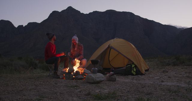 Couple is enjoying a relaxing evening by their campfire near a tent set against mountainous scenery at dusk. Suitable for themes of outdoor adventure, romantic getaways, nature appreciation, camping experiences, and travel promotions.