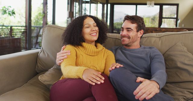 Portrait of happy diverse couple sitting on couch in living room, embracing and laughing. spending free time together at home.