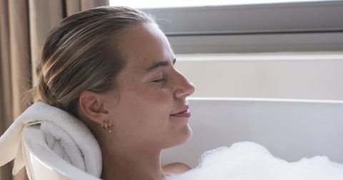 Relaxing woman reclining in bubble bath by window with towel headrest and natural light