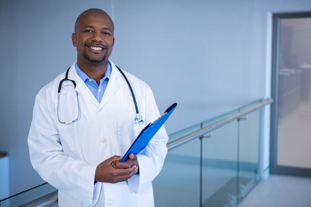 This image depicts a confident male doctor standing in a hospital corridor, holding a clipboard and smiling. Ideal for use in healthcare-related articles, medical websites, hospital brochures, and promotional materials highlighting professional healthcare services.