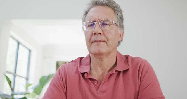 Senior man wearing pink polo shirt and eyeglasses, sitting in a bright room with large windows, looking thoughtful. This image is useful for themes related to aging, wisdom, home life, relaxation, and contemplation. It is ideal for articles, blogs, healthcare advertisements, and social media promoting healthy and active senior living.