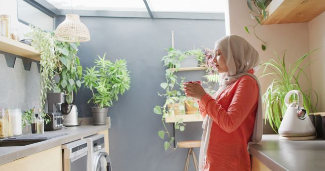 Woman wearing hijab standing next to modern kitchen counter, drinking hot beverage. Walls adorned with various indoor plants, providing a refreshing, green atmosphere. Ideal for use in articles about morning routines, lifestyle, home decor, and promoting relaxation or domestic life.