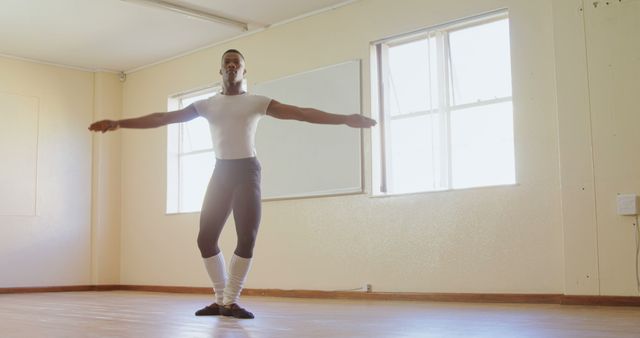 This image shows a male ballet dancer practicing in a well-lit ballet studio. The dancer is executing a balanced pose with arms extended, conveying discipline and grace. Perfect for use in articles about dance training, performing arts, or the dedication required for professional ballet. Can also be used for promoting ballet workshops, fitness regimes associated with dance, or as a visual representation of hard work and determination in performing arts.