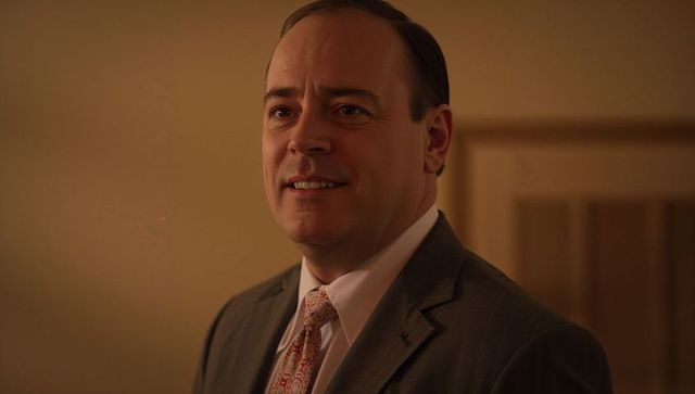 Smiling executive adjusting paisley tie wearing gray suit, white shirt in warm amber light
