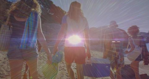 Walking friends carrying cooler and towels on sunlit beach with sunburst flare
