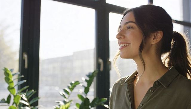 Standing woman gazing through sunlit window with indoor plants, urban home interior