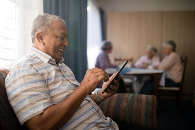 Happy senior man using phone while sitting on sofa by friends at bnursing home