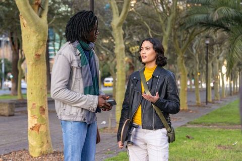 Friends having casual conversation on tree-lined city walkway with camera and smartphone