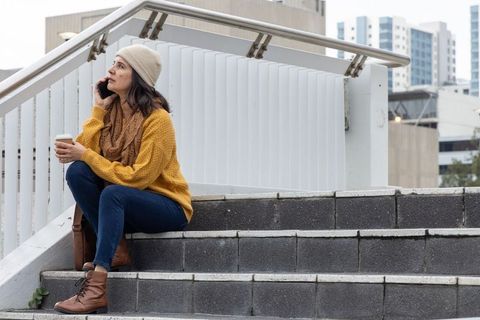 Woman sitting on urban steps talking on smartphone holding takeaway coffee cup commuting