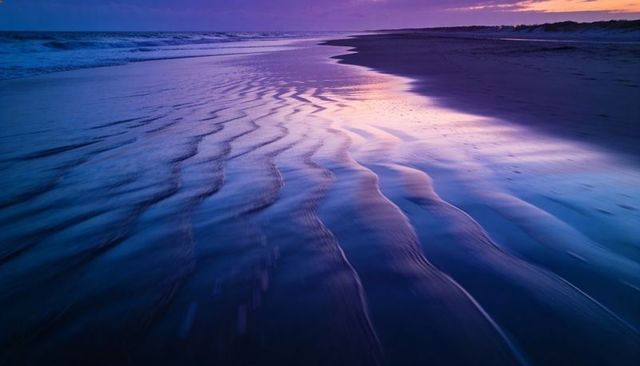 Twilight ripples reflecting wet sand and tidal water on coastal shoreline with purple blue glow