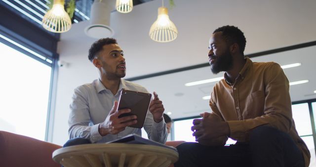 Describing colleagues engaged in a brainstorming session, this image captures two men, one holding a tablet, sitting casually in a modern office lounge area. Ideal for illustrating themes of teamwork, collaboration, and technology in a professional or creative workplace.