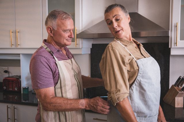 Senior couple spending quality time together in the kitchen. The man is helping the woman with her apron, showcasing their bond and teamwork. Ideal for use in articles or advertisements related to senior living, family life, home activities, and quarantine experiences.