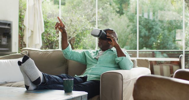 Image depicts a man sitting on a sofa in a living room, using a virtual reality headset while relaxing with his feet up on the table. This can be used to illustrate modern technology in everyday life, concepts of relaxation and entertainment, or technological trends in home environments.
