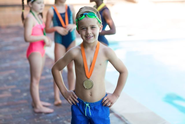 Young boy standing proudly by the poolside, wearing a medal around his neck and swim goggles on his head. Other children in swimwear are visible in the background. Ideal for use in materials related to children's sports, swimming competitions, summer activities, and youth achievements.