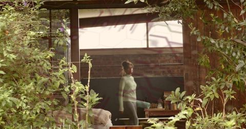 Woman walking with teal yoga mat through rustic sunlit plant-filled studio wearing sweater and leggi