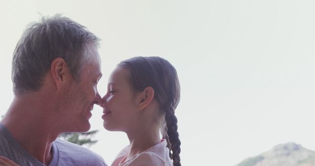 Father holding his daughter while they touch noses, creating a tender and happy moment. They are outdoors during the daytime with a natural, somewhat blurred background, suggesting a peaceful environment. Perfect for family-themed promotions, parenting blogs, advertisements promoting familial affection, and outdoor bonding activities.