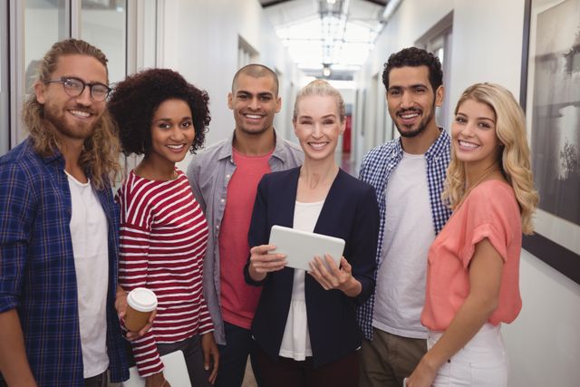 Diverse group of young business professionals standing in an office corridor, smiling and holding a tablet and coffee cup. Ideal for use in articles or advertisements about teamwork, collaboration, modern workplaces, and diversity in the workplace.