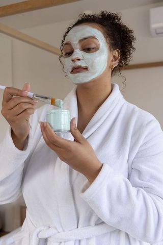 African american woman applying mint facial mask wearing white bathrobe at home