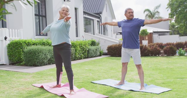 This image shows a senior couple practicing yoga outdoors in their home's garden. They are standing on yoga mats with their arms outstretched, enjoying a sunny day. This visual is perfect for promoting fitness, wellness, and active lifestyle among seniors. Ideal for use in health and wellness websites, retirement living brochures, and fitness campaigns targeting elderly populations.