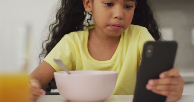 Young girl with curly hair wearing yellow shirt, eating breakfast and looking at smartphone. Ideal for educational content on balancing technology use with healthy eating. Useful for illustrating modern family life, childhood habits, or health and nutrition topics.