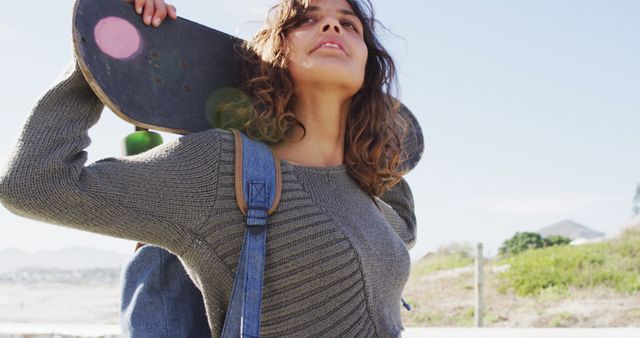 Biracial woman holding skateboard behind head standing in the sun enjoying the view by the sea. healthy living, off the grid and close to nature.