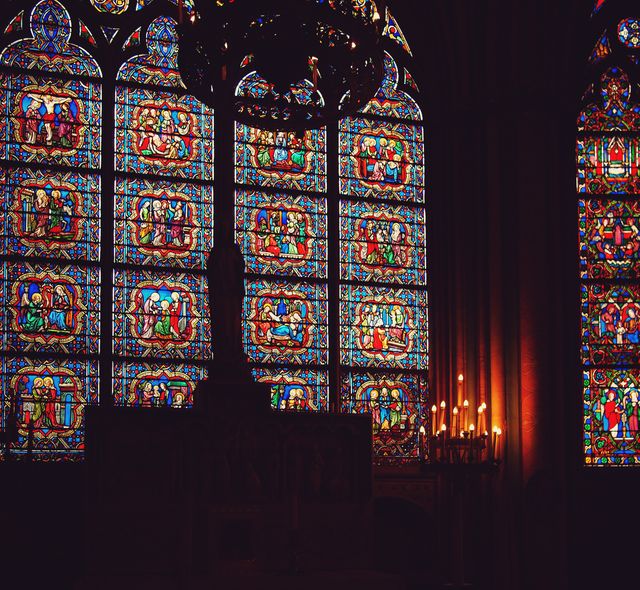 Detailed stained glass windows provide a bright and colorful display in a dimly lit church interior. Lit candles add warmth and a sense of spirituality to the scene. Perfect for use in articles or content about religious practices, history of stained glass art, or architectural beauty of churches.