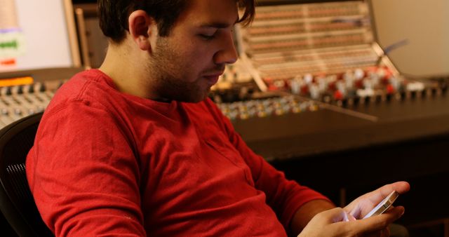 Young man wearing red shirt using smartphone in a professional audio or music studio control room. Surrounded by mixing equipment and consoles, the man focuses intently on his phone. Useful for themes relating to technology, music production, modern workplaces, or personal and professional communication.