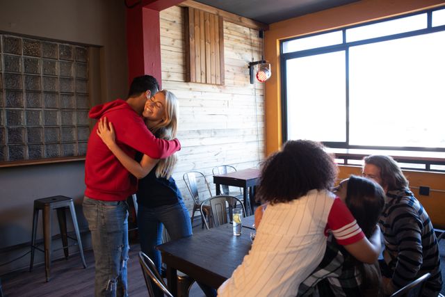 Group of multi-ethnic male and female friends at the bar in a pub during the day, man and woman greeting, hugging and smiling socialising together. Friendship leisure time fun.